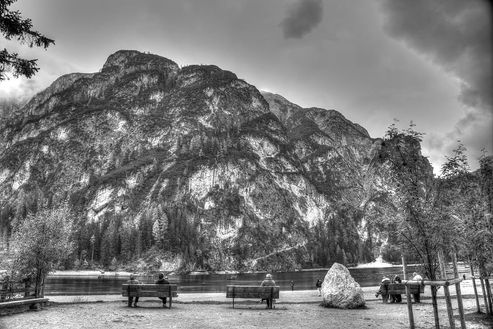 Pessoas sentadas em bancos de madeira à beira de um lago, de frente para uma grande montanha de rocha.