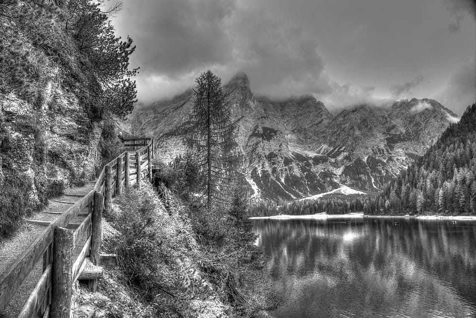 Passadiço de madeira encostado à rocha ao longo de um lago calmo que reflete as montanhas e a floresta.
