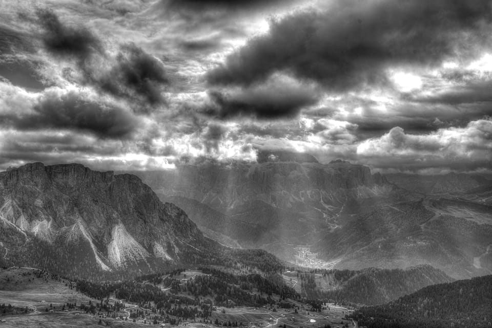 Vale entre montanhas de rocha, com feixes de luz a atravessar nuvens escuras e uma vila pequena no fundo.